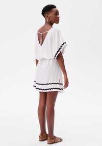 Witte jurk met zwart zigzag accent, lichtgewicht stof, V-rug ontwerp en een cinched taille. Model draagt platte sandalen en oorbellen met een ring.