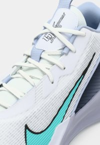 Zapatilla deportiva blanca y azul claro con cordones blancos, logotipo en turquesa y negro de swoosh, y la marca "Academy" en la lengüeta.