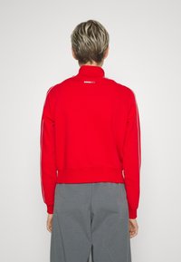 Röd sweatshirt med hög krage, vita sidostråk och ribbade mudd. Har en liten logotyp på baksidan. Matchas med grå byxor.