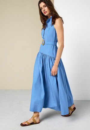 Frau trägt ein ärmelloses blaues Maxikleid mit Gürtel und hellbraunen Sandalen, steht mit einem Fuß nach vorne gegen einen neutralen Hintergrund.