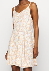 Robe d'été fleurie à fines bretelles. Présente une palette de couleurs pastel, principalement pêche et blanc, avec des motifs floraux et une silhouette évasée.