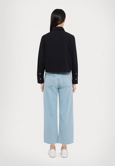 Cropped shirt nera con polsini abbottonati, abbinata a jeans a gamba larga azzurri e sneakers bianche, con dettagli di orlo sfrangiato.