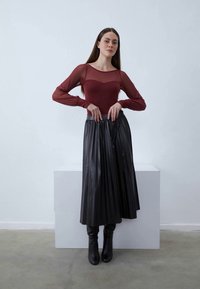 Burgundy hosszú ujjú hálós felső, fekete plissé bőr szoknya és térd fölé érő fekete csizmával párosítva, minimalista háttér előtt.