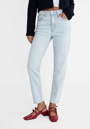 Person iført lyseblå højtaljede slanke jeans, mørk sweater og røde Mary Jane-sko med lav hæl, stående med den ene hånd i lommen.