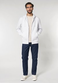 Felpa bianca con zip e tasche anteriori, abbinata a una maglietta beige chiaro, jeans denim blu scuro e sneakers bianche. Vestibilità casual.