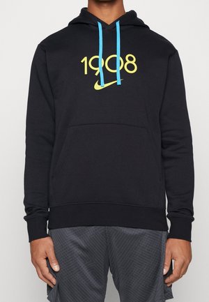 Osoba ubrana w czarną bluzę z kapturem z jasnoniebieskimi sznurkami i żółtym napisem "1908" oraz logo Nike na przodzie, w połączeniu z ciemnymi, wzorzystymi szortami.