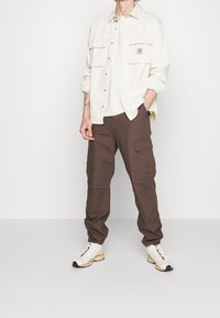 Conjunto de hombre que incluye una camisa crema de botones con dos bolsillos en el pecho, pantalones cargo marrones y zapatillas blancas con acentos multicolores.
