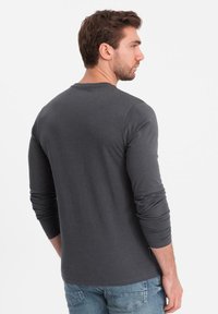 Man med brunt hår som bär en mörkgrå långärmad t-shirt och blå jeans, vänd åt sidan mot en enkel vit bakgrund.