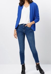 Blauer strukturierter Cardigan über einem weißen V-Ausschnitt-Top, kombiniert mit eng anliegenden dunkelblauen Jeans und schwarzen Ankle Boots. Das Outfit hat eine lässige Passform.