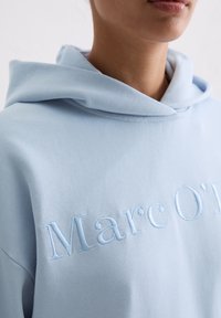 Zbliżenie osoby w jasno-niebieskiej bluzie z kapturem z podniesionym białym haftowanym napisem na piersi, ukazujące część ust i szyi.