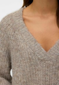 Pull en tricot marron clair avec un col en V. La texture est douce et duveteuse, avec une coupe ample et des poignets côtelés.