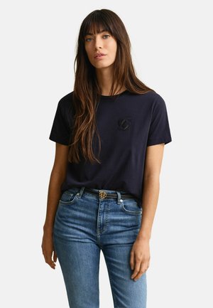 Vrouw met lang bruin haar, gekleed in een zwart T-shirt met korte mouwen met een klein logo en een blauwe spijkerbroek met een zwarte riem.