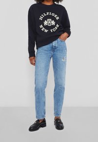 Marinblå sweatshirt med vit text "HILFIGER NEW YORK", ljusblå slitna jeans och svarta loafers med guldaccenter. Avslappnad outfit.