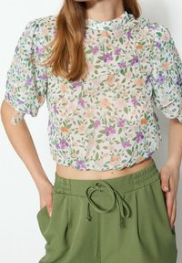 Blusa cropped floreale in tessuto trasparente bianco con motivi arancioni, viola e verdi. Maniche a sbuffo e scollatura tonda. Abbinata a pantaloni verdi.