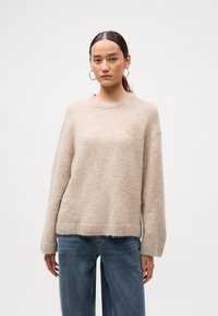 Béžový pletený sveter s voľným strihom, guľatým výstrihom a rebrovanými manžetami. Textúrovaná tkanina s jemným, nadýchaným vzhľadom. Nosený s modrými džínsami.