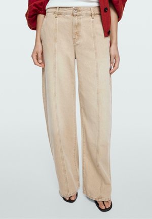 Person iført beige bukser med vide ben og synlige forreste sømme, sorte åbne sandaler og delvist udsyn til en rød jakke og hvid top.