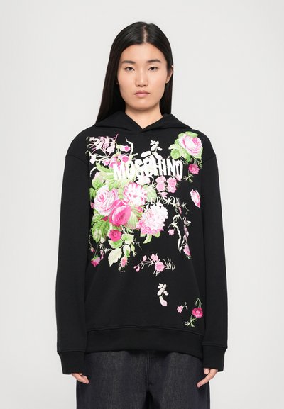Hoodie noir arborant des graphiques floraux éclatants en rose et vert, avec un design de texte "Moschino" bien visible. Coupe douce et décontractée.
