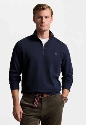 Sudadera de media cremallera azul marino oscuro con puños acanalados, que presenta un pequeño logotipo en el pecho y un cuello en contraste. Combinada con pantalones de pana marrones.