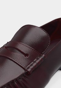 Γυαλιστερό μπορντό δερμάτινο μοκασίνι με λουράκι για κέρματα και λεπτομερή ραφή κατά μήκος του ποδόγυρου και των πλευρών.