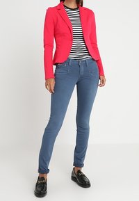 Blazer rose cintré porté sur une chemise rayée noir et blanc, associé à un jean slim bleu et des mocassins noirs. Textures lisses et design sur mesure.