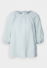 Blouse bleu clair avec un motif texturé à carreaux, des manches bouffantes élastiquées et une fermeture en forme de trou de serrure à l'arrière.