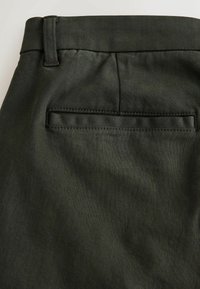 Dunkelgrüne Hose mit glatter Textur, gerader Form, Gürtelschlaufen und einer einzigen Gesäßtasche mit gesticktem Randdetail.