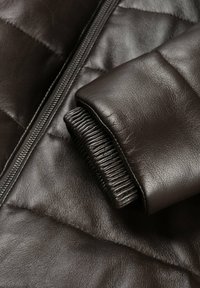 Brun læderjakke med en quiltet design, teksturerede manchetter og en frontlynlås. Detaljerne inkluderer ribstrik ved ærmeåbningen.