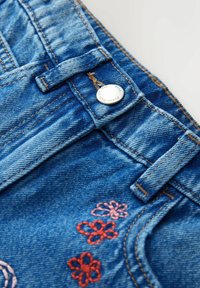 Détail d'un jean en denim bleu avec un bouton en métal, des passants de ceinture, des fleurs brodées orange et roses près de la couture de la poche.