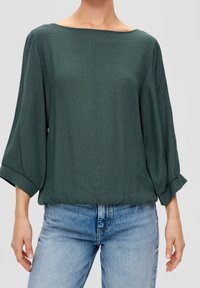 Blusa de color verde oscuro holgada con mangas tres cuartos, tela texturizada y escote redondo amplio, combinada con jeans de lavado claro.