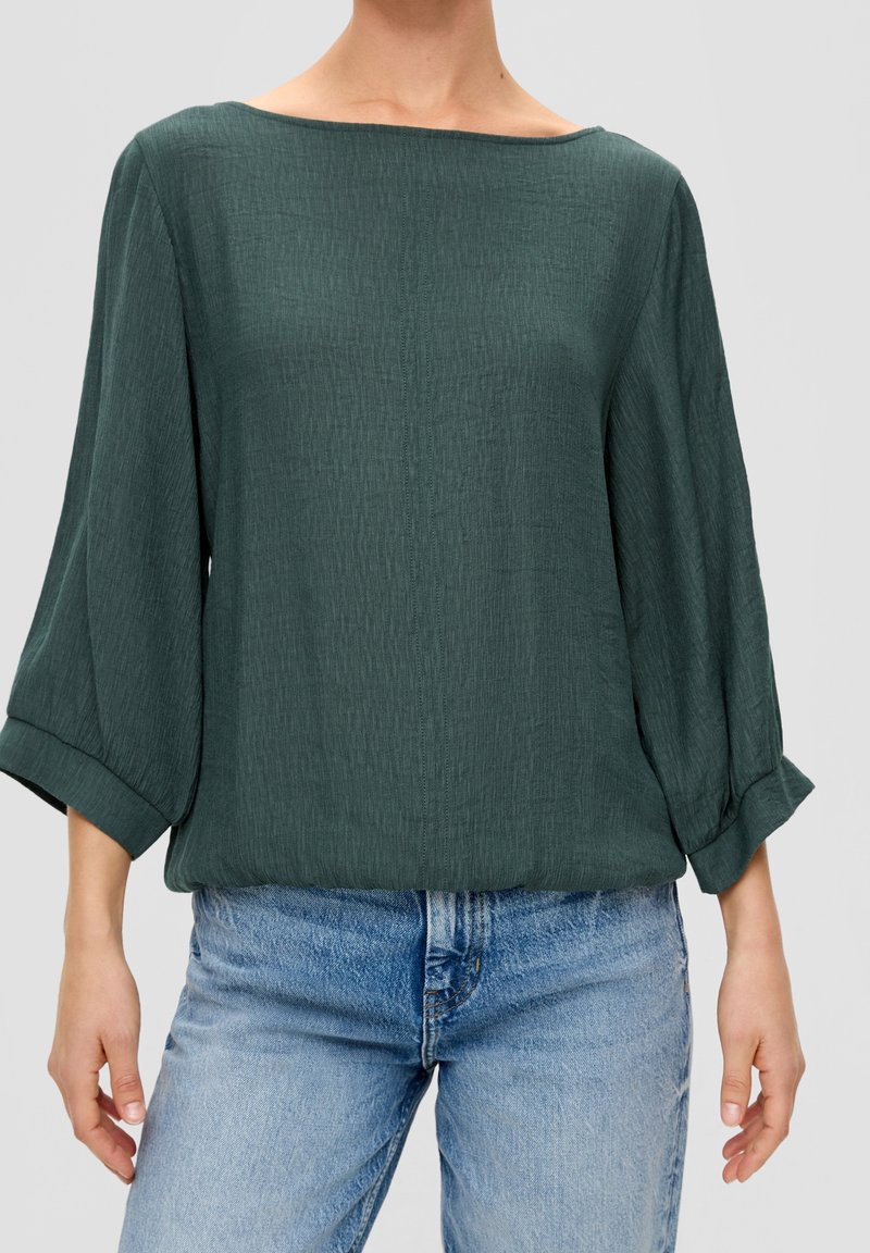 Blusa de color verde oscuro holgada con mangas tres cuartos, tela texturizada y escote redondo amplio, combinada con jeans de lavado claro.