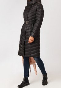 Giacca a puff nera con vita con cintura, colletto alto e motivo trapuntato. Indossata sopra jeans scuri e stivaletti neri.