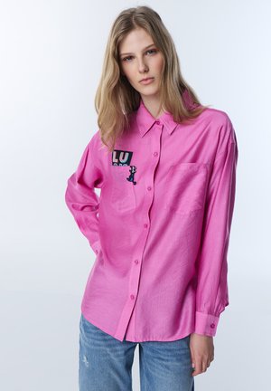 Młoda kobieta o długich blond włosach ma na sobie różową koszulę sztruksową z naszywkami oraz niebieskie dżinsy, pozuje na jednolitym białym tle.