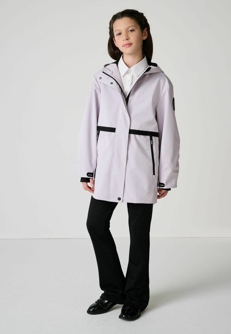 Svetlofialová pršiplášť s čiernymi akcentmi, predný zips, nastaviteľná kapucňa a dve vrecká na zips v kombinácii s čiernymi nohavicami a topánkami.