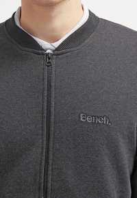 Gros plan sur une personne portant un sweat-shirt à fermeture éclair gris foncé avec "Bench." brodé sur le côté gauche de la poitrine, par-dessus une chemise à col gris clair.