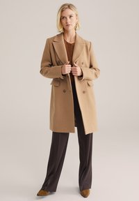 Cappotto doppiopetto in misto lana color cammello con rever e tasche a pattina, abbinato a pantaloni a gamba larga marrone scuro e tacchi marroni.