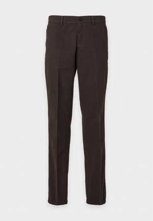 Un homme porte une veste marron, une chemise rayée, un pantalon foncé et des bottes. Il tient un sac fourre-tout kaki sur un fond uni et clair.