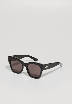 Óculos de sol pretos com uma armação quadrada robusta, lentes escuras e a marca "GUCCI" em dourado na haste. Acabamento em plástico suave e superfície mate.