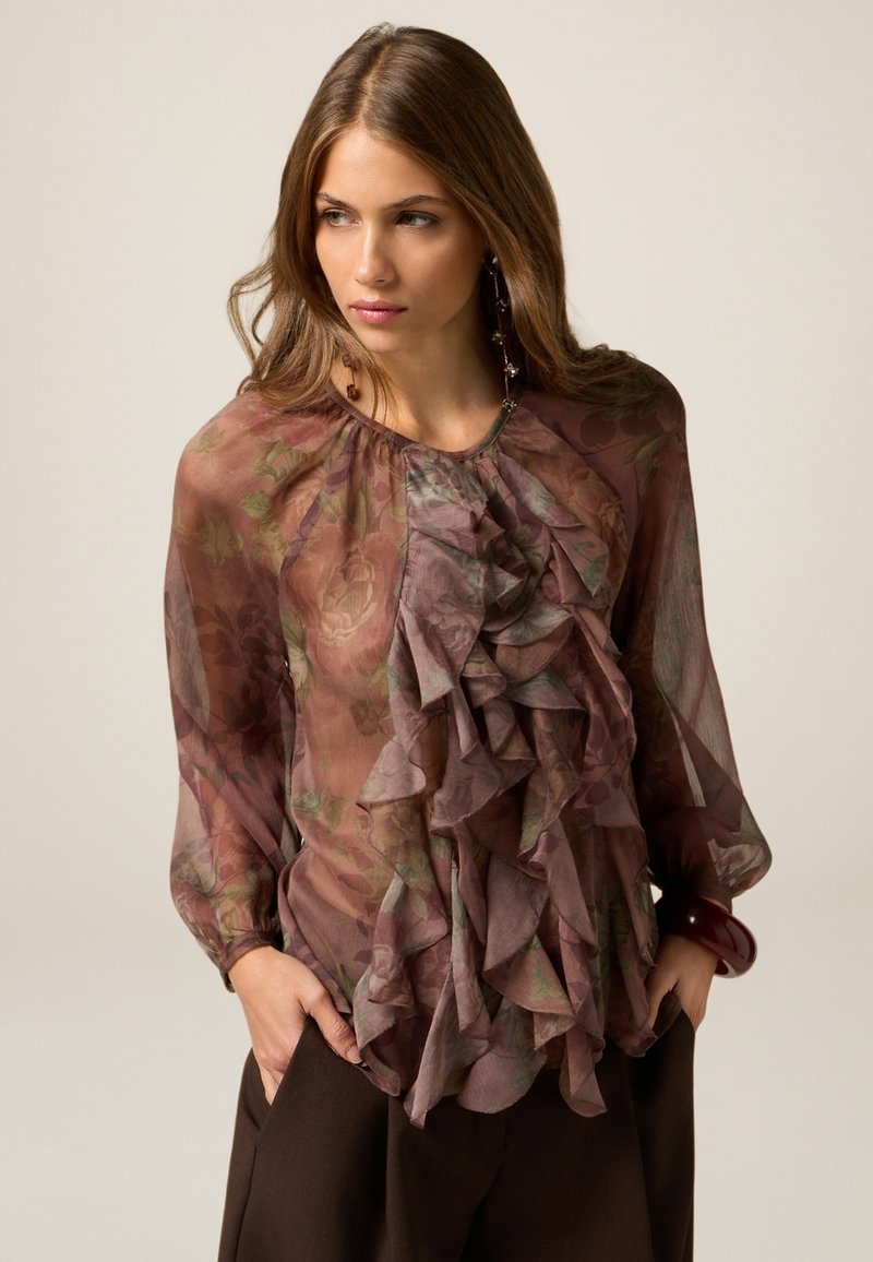 Durchsichtige, mauvefarbene Bluse mit Rüschendetails, floralen Mustern und langen Ärmeln. Kombiniert mit dunkelbraunen Hosen, die einen schlichten Ausschnitt haben.