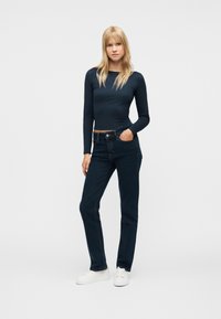 Γυναίκα με ξανθά μαλλιά, φοράει εφαρμοστή μπλε σκούρη μπλούζα μακρυμάνικη, σκούρο μπλε τζιν, λευκά αθλητικά παπούτσια, στέκεται με το ένα χέρι στην τσέπη μπροστά σε απλό φόντο.