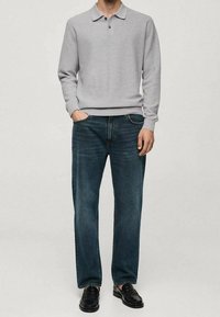 Mann trägt ein hellgraues Langarm-Poloshirt, dunkelblaue Jeans und schwarze Lederloafer, steht mit einer Hand in der Tasche.