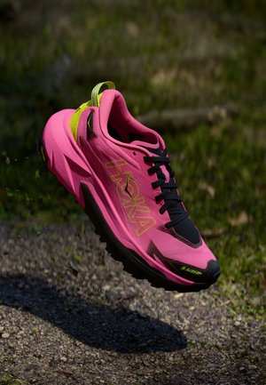 Zapatilla de running Hoka con una parte superior de malla rosa, cordones negros y detalles verdes. Presenta una suela gruesa con un diseño de taco agresivo para mayor tracción.