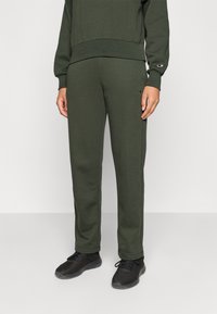 Pantaloni de trening verde măsliniu cu talie elastică, buzunare laterale și logo brodat pe buzunarul din stânga. Materialul pare moale și elastic.