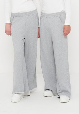 Due persone che indossano pantaloni larghi grigio chiaro con bottoni a pressione laterali e sneakers bianche, in piedi davanti a uno sfondo bianco semplice.