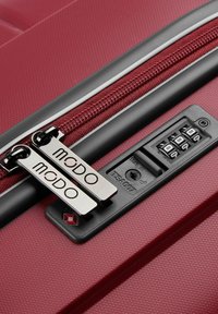 Maleta dura roja con superficie texturizada, doble cremallera plateada con etiquetas de marca y un mecanismo de bloqueo aprobado por la TSA en negro.