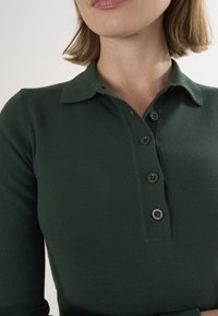 Mørkegrøn strikket polo med krave, fem sorte knapper og figursyede ærmer, med et tekstureret stof og et strømlinet design.