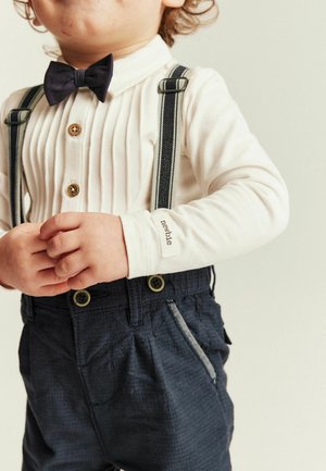 Toddler wearing a white buttoned shirt with black bow tie, striped suspenders, and dark blue pants, hands clasped in front.