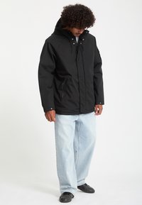 Homme aux cheveux bouclés portant une veste noire à capuche, un jean bleu clair ample et des chaussures noires, debout la tête baissée.