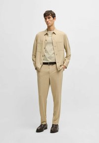 Giacca camicia beige a trama sopra una t-shirt beige chiaro, abbinata a pantaloni coordinati. Scarpe marroni completano l'outfit.