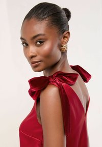 Piros selyemruha bársonyos masnikkal a vállakon, mély V kivágással és sima textúrával. Modell arany spirál fülbevalóval.