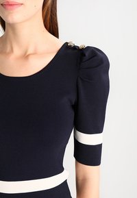 Femme portant un haut bleu marine avec des manches bouffantes, des rayures horizontales blanches sur les manches et la taille, et des boutons dorés sur l'épaule.